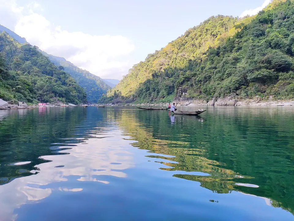 Green hills mirrored in the glassy Umngot River at Dawki, with a lone wooden boat mid-stream