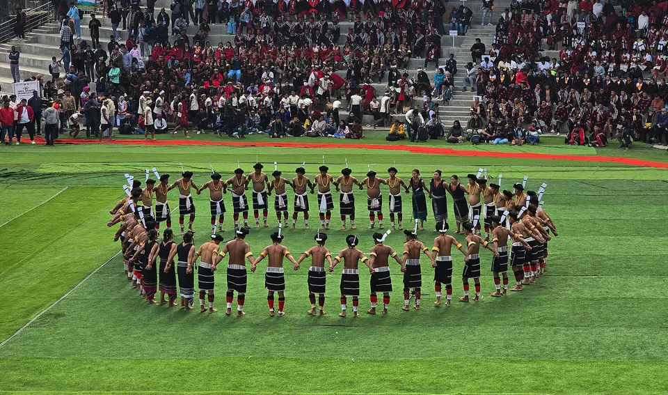 Naga dancers in a wide circle on the arena lawn at the Hornbill Festival, with packed stands behind