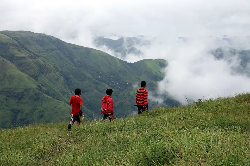Three children in red walking along a grassy ridge as clouds roll through the valleys near Sohra